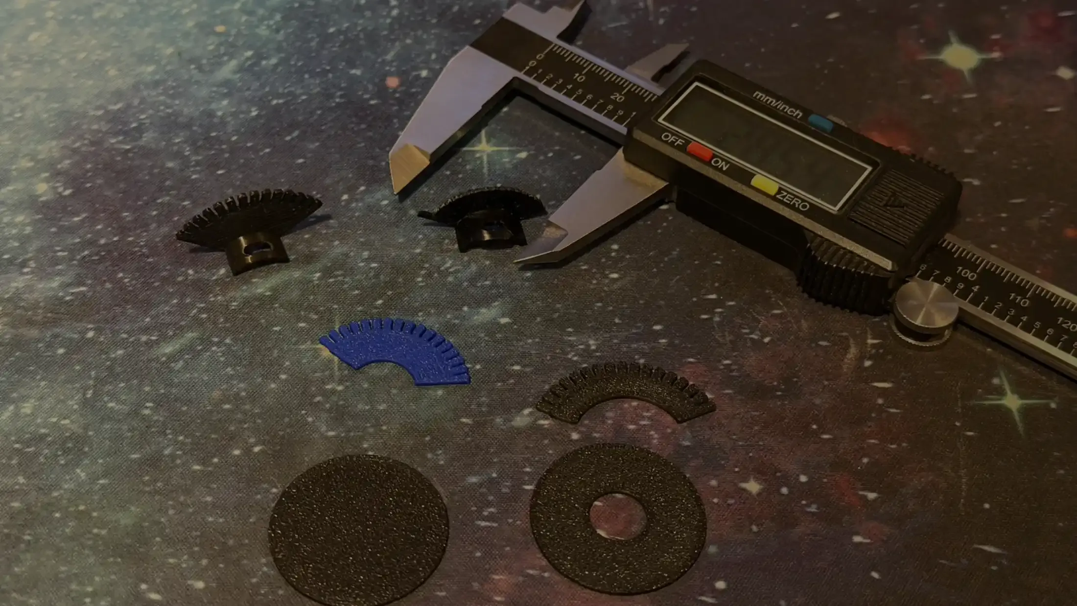 Digital calipers beside a row of 3D-printed replacement gear parts in black and blue, from early attempts to the working part